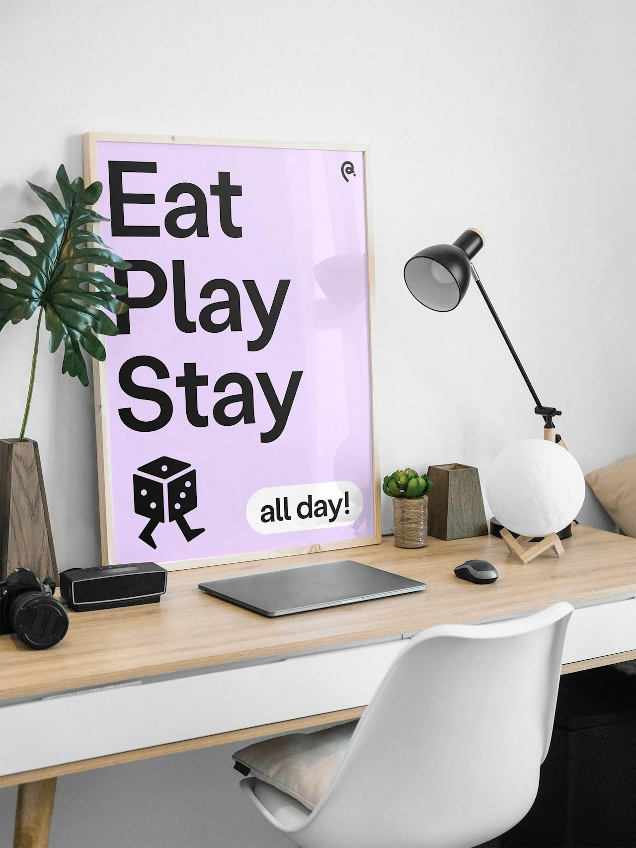 Juliste puupöydällä, jossa teksti "Eat Play Stay all day!" Sisältää noppakuvituksen, ympärillä toimistotarvikkeita kuten kasvi, lamppu ja kannettava tietokone minimalistisessa huoneessa.