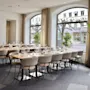 A long dining table is set with glasses and cutlery, surrounded by beige chairs, in a well-lit room with large windows and street view outside. Text: "SOC".