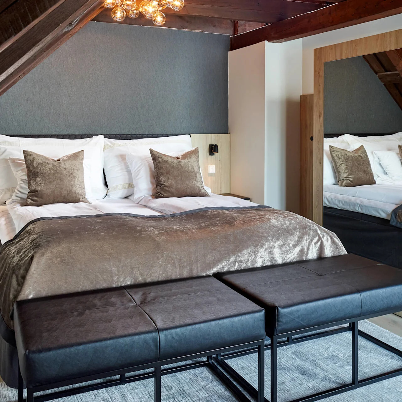 A well-made bed with brown bedding in a cozy, wood-accented bedroom with sloped ceilings and a reflective chandelier.