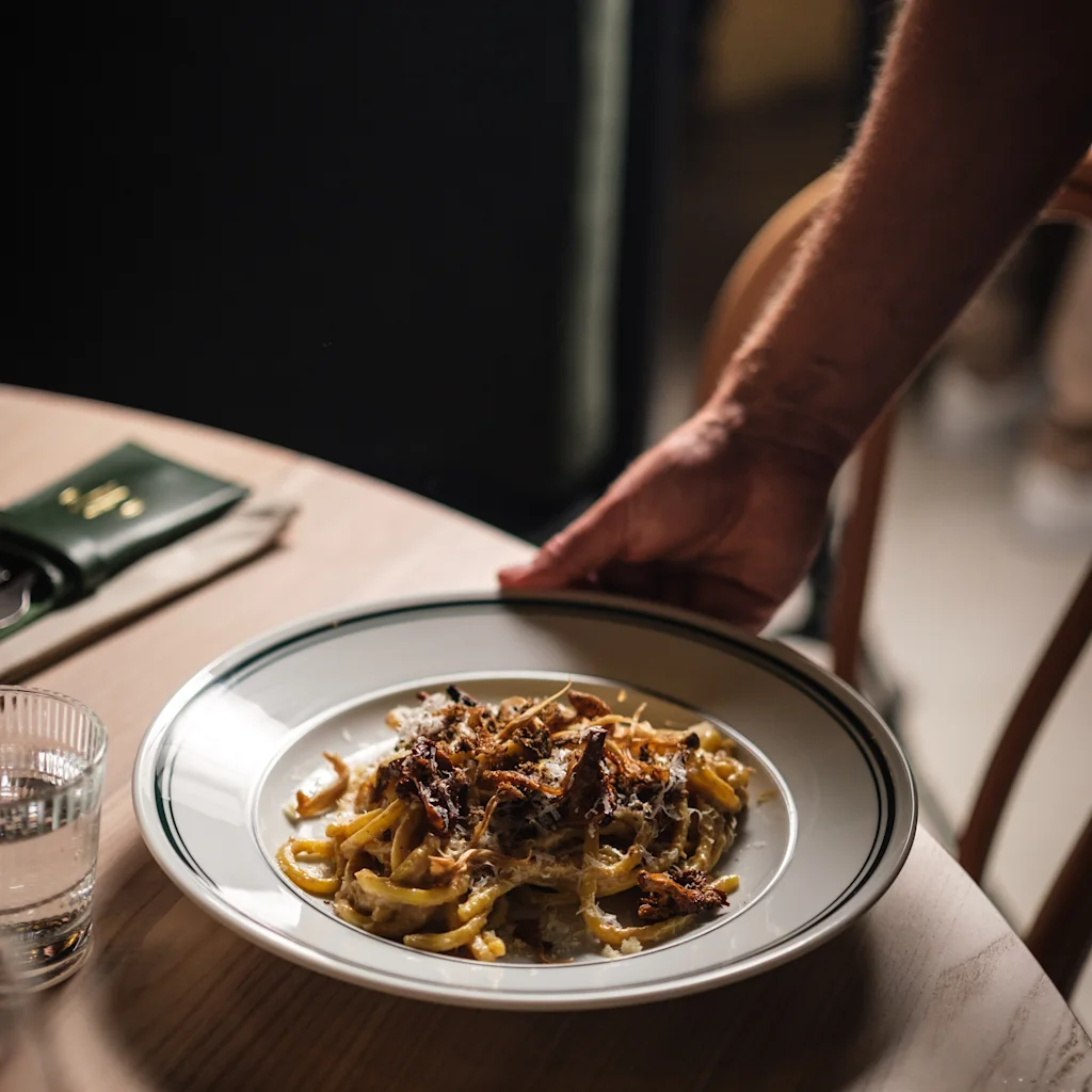 En persons hånd serverer en deilig tallerken pasta ved et restaurantbord, med et glass vann i nærheten.