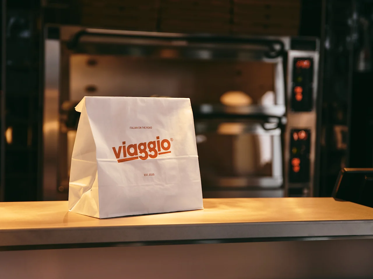 En hvit papirpose, merket "ITALIAN ON THE ROAD EST. 2025", hviler på en disk på et hotellkjøkken med en ovn.