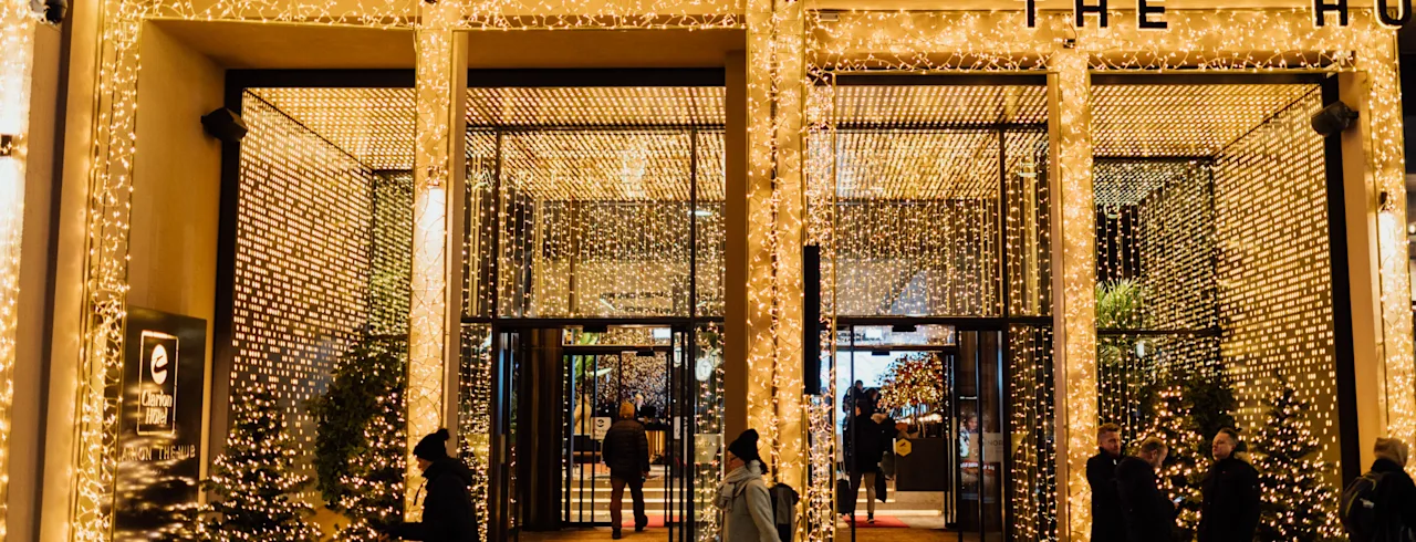 People walk in front of a brightly lit hotel entrance, adorned with Christmas lights, creating a festive atmosphere at night.