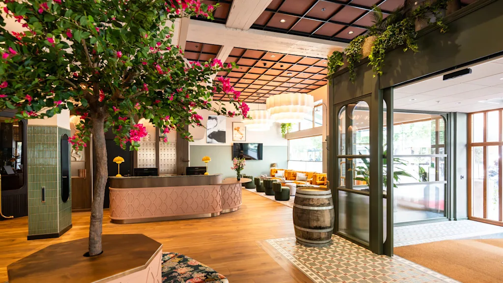 A large tree with pink flowers decorates a stylish hotel lobby featuring a wooden floor, green tiled walls, a reception desk, and cozy seating area. A decorative barrel and plants complete the setting.