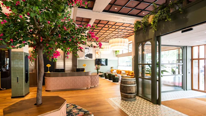 Et stort tre med rosa blomster pynter en stilfull hotellobby med tregulv, grønne flisevegger, en resepsjonsdisk og koselig sitteområde. En dekorativ tønne og planter fullfører settingen.