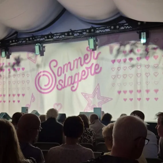 Et publikum ser på en scene med et bakteppe som sier Sommerslagere, dekorert med rosa hjerter og en stjerne.