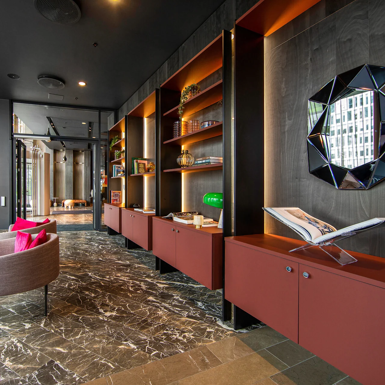 Elegant lounge features modern chairs with pink cushions beside a marble table; shelves displaying books and decor; reflective geometric mirror on wall; large open doorway extends to another room.