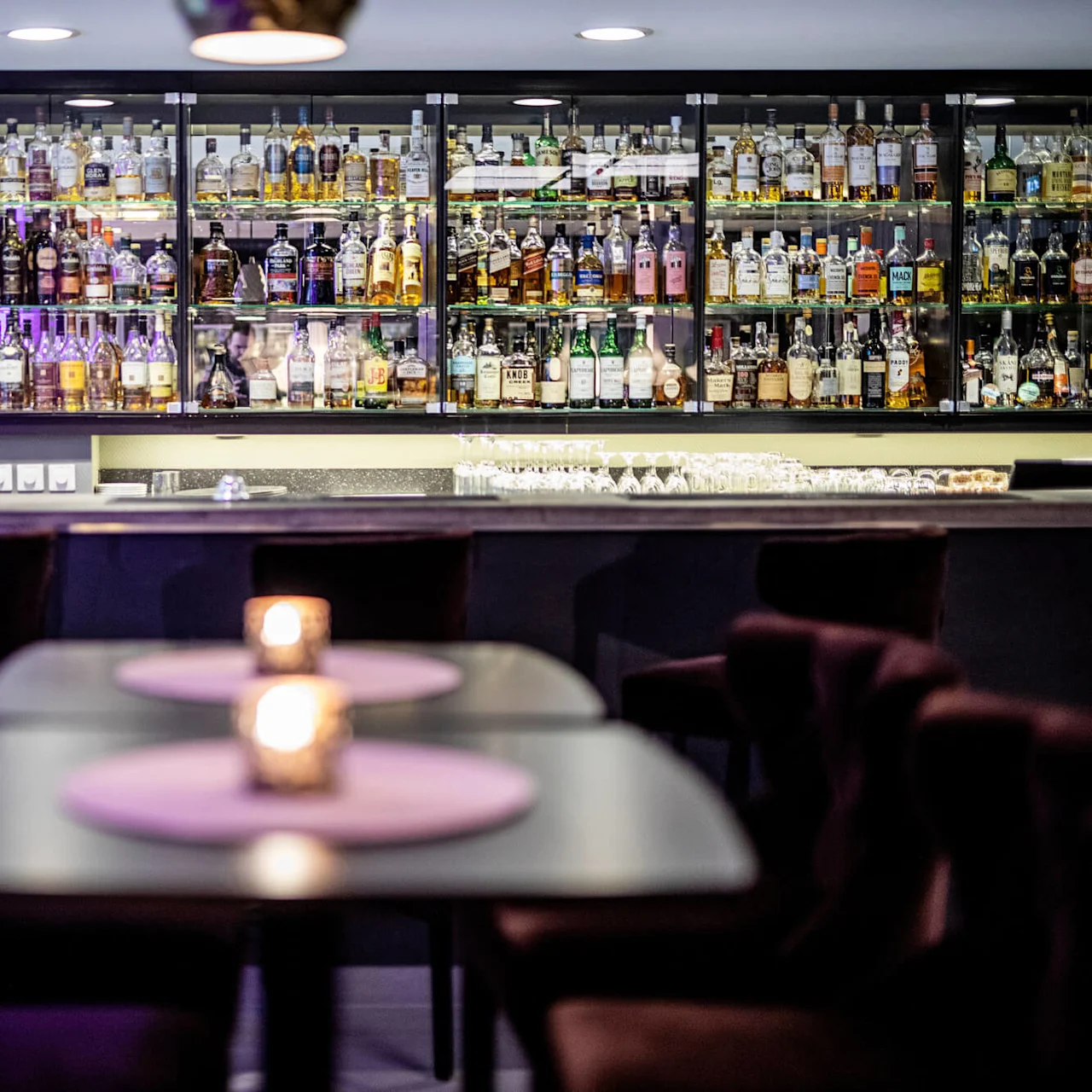 A well-stocked bar glows with purple lighting, bottles lined up, foreground showing candlelit tables, and luxurious chairs in a lounge.