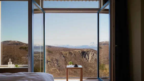 Open window frames a stunning mountain view; a wooden table holds a cup and plant. Inside, part of a bed with white sheets is visible.