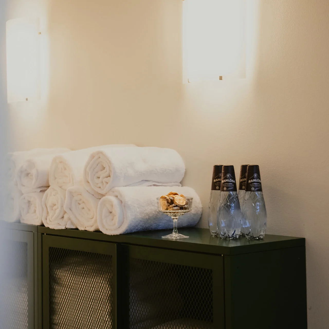 White towels are neatly rolled and stacked on a dark cabinet beneath wall lights. A glass bowl with snacks and three bottles labeled "Ramlösa" are positioned nearby.