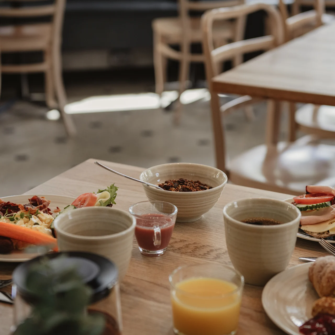 Frukostbord med tallrikar av mat och kaffekoppar på ett träbord; tomma stolar syns i bakgrunden, vilket skapar en mysig kafémiljö.