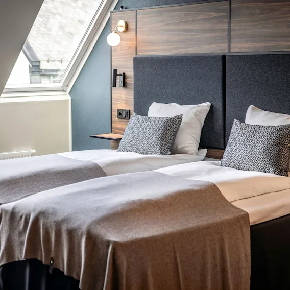 Moderne hotellrom med to senger, mønstrede puter, brune pledd, mørke sengegavler og et takvindu.