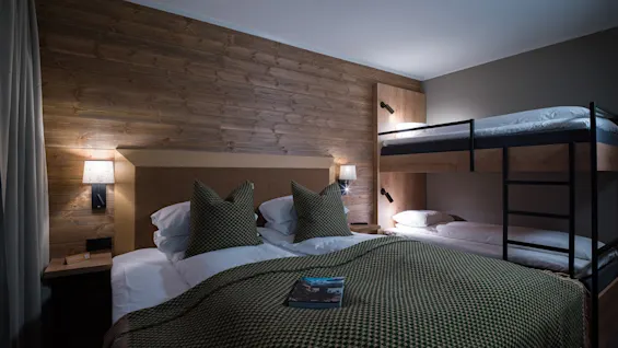 A neatly made bed with green checkered bedding and pillows, next to a wooden wall, under two modern lamps; a bunk bed is beside it in a cozy room.