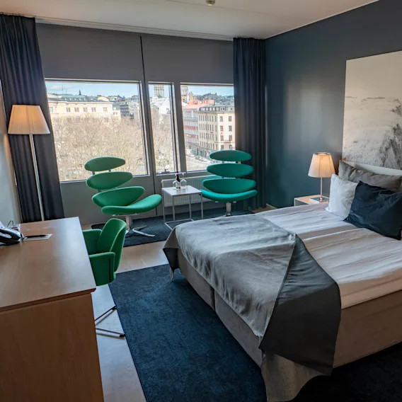 En prydligt bäddad säng med grått sängkläder står mot en mörk vägg med ett stort kustfotografi. Gröna stolar och ett litet bord vid fönstret ger utsikt över stadslandskap.