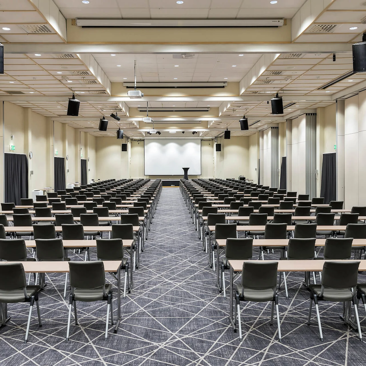 Rader av tomma stolar och bord vetter mot en stor projektionsskärm i ett rymligt, välbelyst konferensrum. Taket har hängande högtalare och lampor. Gröna utgångsskyltar är synliga.