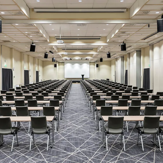 Rows of empty chairs and tables face a large projection screen in a spacious, well-lit conference room. The ceiling features hanging speakers and lights. Green exit signs are visible.