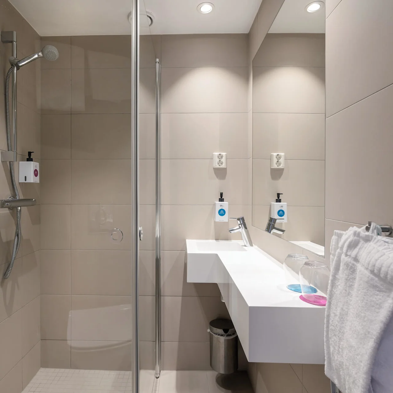 Et moderne bad har en glasslukket dusj, veggmontert dusjhode, hvit vask med to dispensere, en håndklestang med et håndkle og overlys. Veggene er flislagt i beige, og det er en søppelbøtte under vasken.