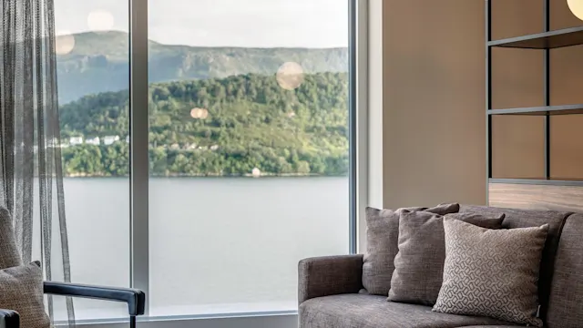 A cozy, modern living room with a gray sofa and round wooden table; large window revealing a scenic lake and mountain view; illuminated by two hanging globe lights.