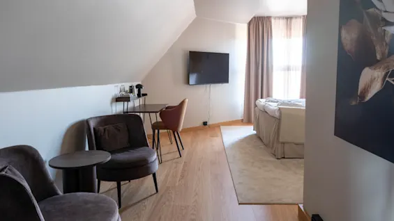 Ljust hotellrum med snedtak, bekväma fåtöljer, ett litet skrivbord, väggmonterad tv och en säng vid fönstret