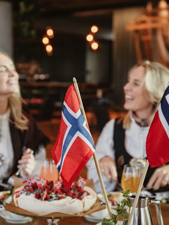 To kvinner i bunad smiler ved et festdekket bord med norske flagg og en pavlova.