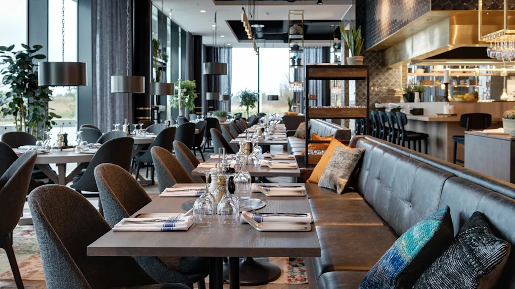 En modern restaurang har snyggt arrangerade matbord med eleganta uppsättningar i ett rymligt rum. Stora fönster ger naturligt ljus, medan dekorativa växter och stiliga möbler skapar en mysig atmosfär.