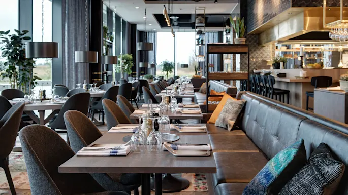 A modern restaurant features neatly arranged dining tables with elegant settings in a spacious room. Large windows provide natural light, while decorative plants and stylish furnishings create a cozy atmosphere.