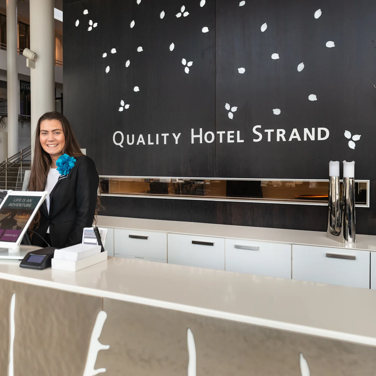 Receptionist smiles behind a desk in a hotel lobby with black wall and white leaf designs. Text reads: "QUALITY HOTEL STRAND" and "LIFE IS AN ADVENTURE."