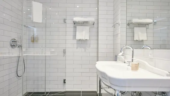 Et rent, moderne badeværelse med en glasbruser, hvide flisevægge og en håndvask med spejl.