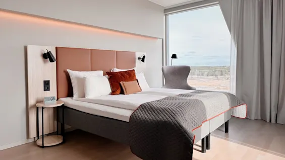 A modern hotel room features a double bed with layered pillows and a quilt, flanked by wall lamps. Large windows offer a scenic view of a forested landscape. 