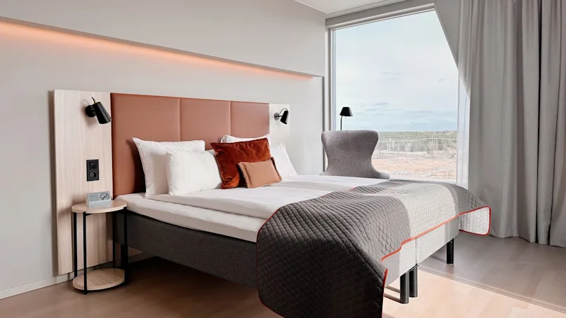 A modern hotel room features a double bed with layered pillows and a quilt, flanked by wall lamps. Large windows offer a scenic view of a forested landscape. 