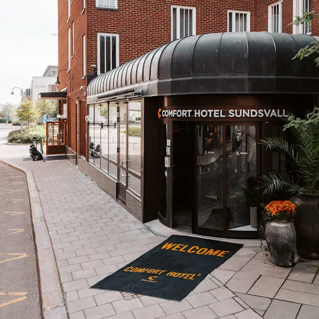 Inngang til Comfort Hotel Sundsvall med sort markise og potteplanter; en velkomstmatte har påskriften "WELCOME COMFORT HOTEL."