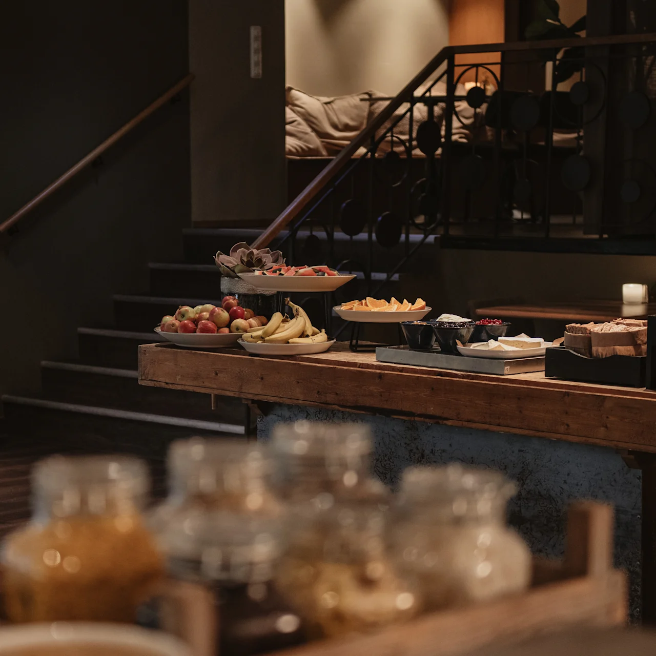 A wooden table holds assorted fruits, bread, and jars under a warm lamp, placed in a dimly lit room with stairs leading up, creating a cozy dining setting.