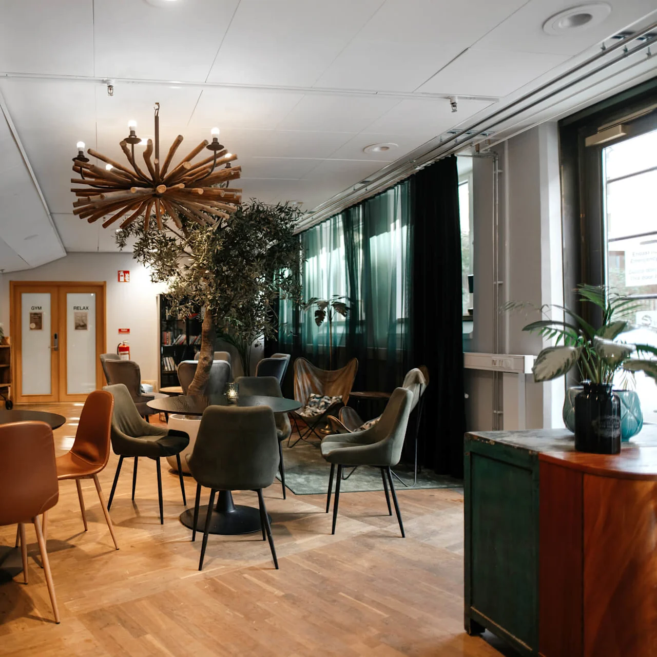 Modern lounge with varied seating and decorative plants, large unique chandelier dominates the space, signs for 'GYM' and 'RELAX' on doors.