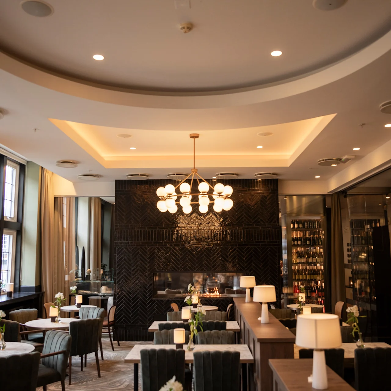 Elegant dining tables set with chairs are placed in a warmly lit, modern restaurant. A chandelier hangs from the ceiling. Large windows with curtains line one wall.