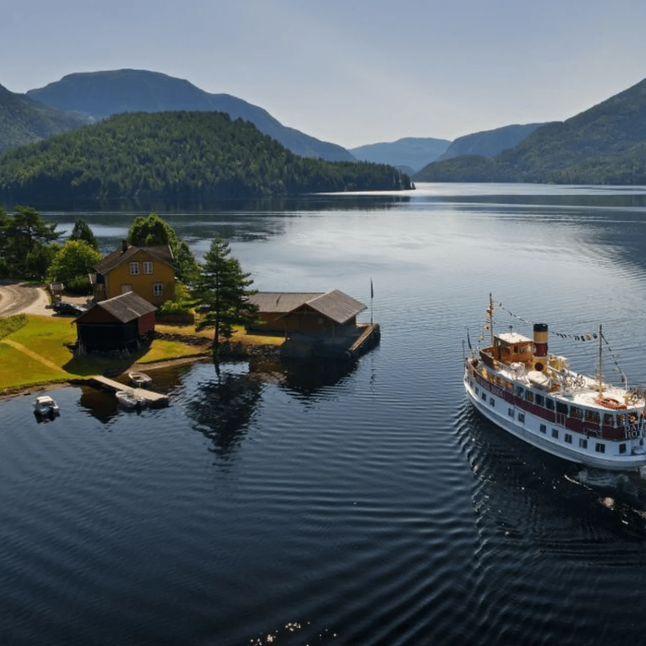 A steamboat sails through a tranquil lake surrounded by lush, forested hills. Near the shore, there's a rustic wooden house and boathouse with small boats docked.