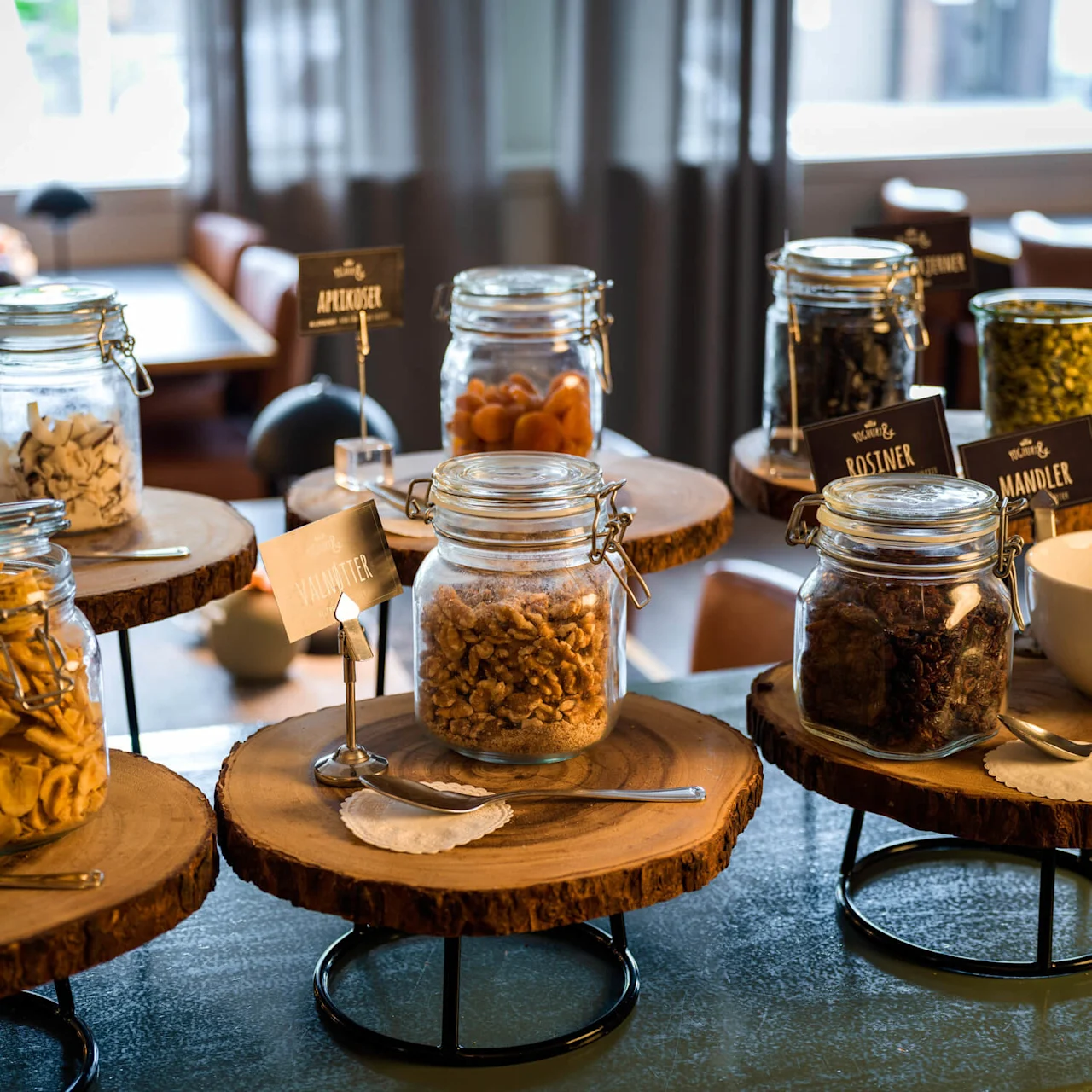 Jars of various dried fruits and nuts are displayed on wooden stands in a dining area. Text: Kokosbiter, Tørket banan, Aprikoser, Valnøtter, Rosiner, Mandler.