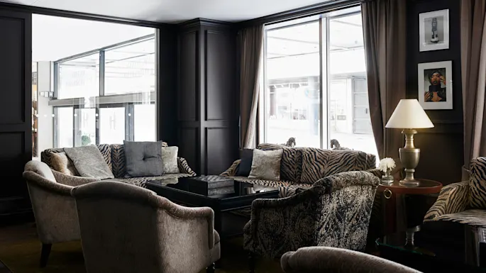 Elegant loungeområde med mönstrade soffor och fåtöljer centrerade runt ett blankt svart bord. Mjuk belysning inkluderar en lampa på ett sidobord nära stora fönster.