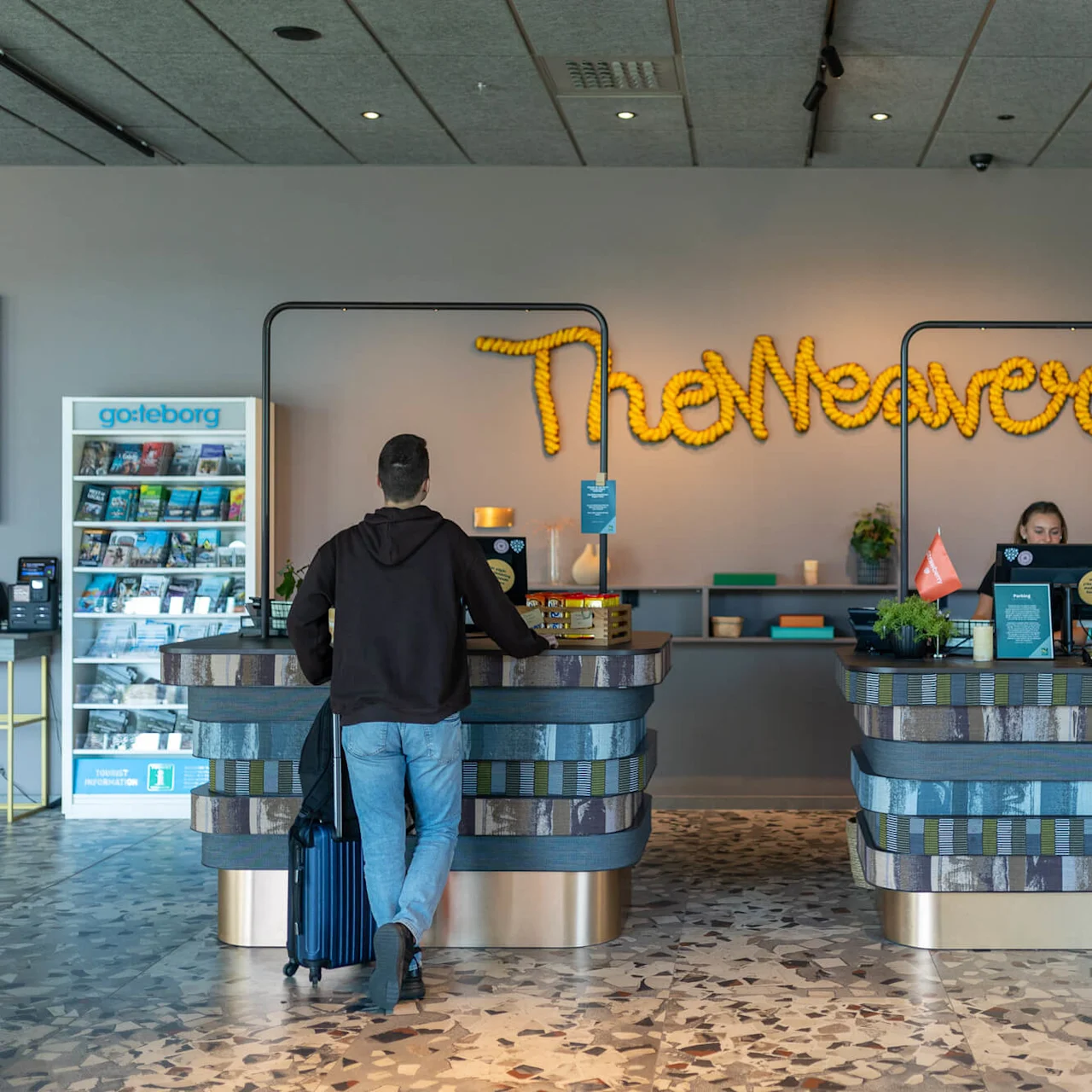 En man med en resväska närmar sig en hotellreception. Bakom disken assisterar en anställd, med texten "The Weaver" på väggen. Broschyrer och växter dekorerar rummet.