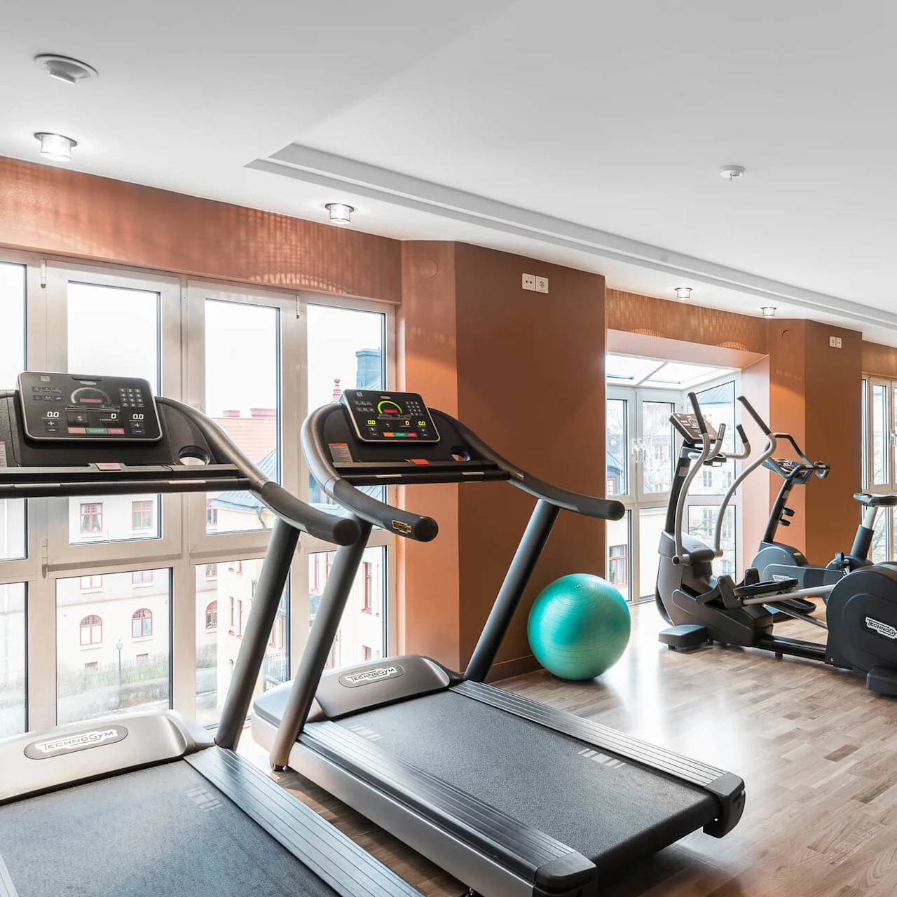Löpband och motionscyklar står i ett ljust gym, reflekterade i speglar från golv till tak, tillsammans med balansbollar och yogamattor på polerat trägolv.
