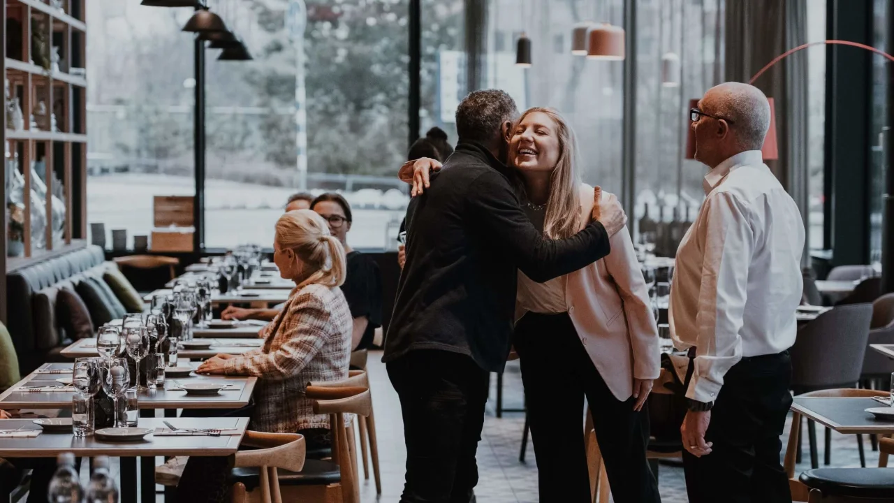 To mennesker omfavner hinanden lykkeligt i en restaurant med siddepladser ved borde.