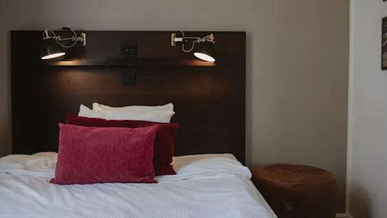 En seng med røde og hvite puter står mot en mørk trehodegavl med leselamper, i et minimalistisk innredet rom med beige vegger.