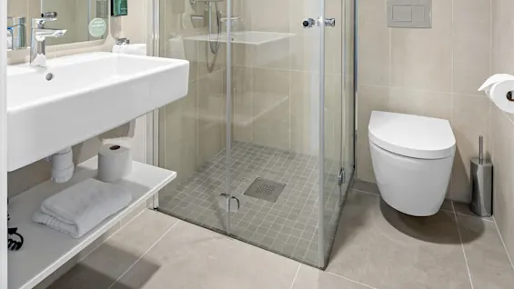 A modern bathroom with beige tiles, featuring a glass shower, sink, and toilet under bright lights.
