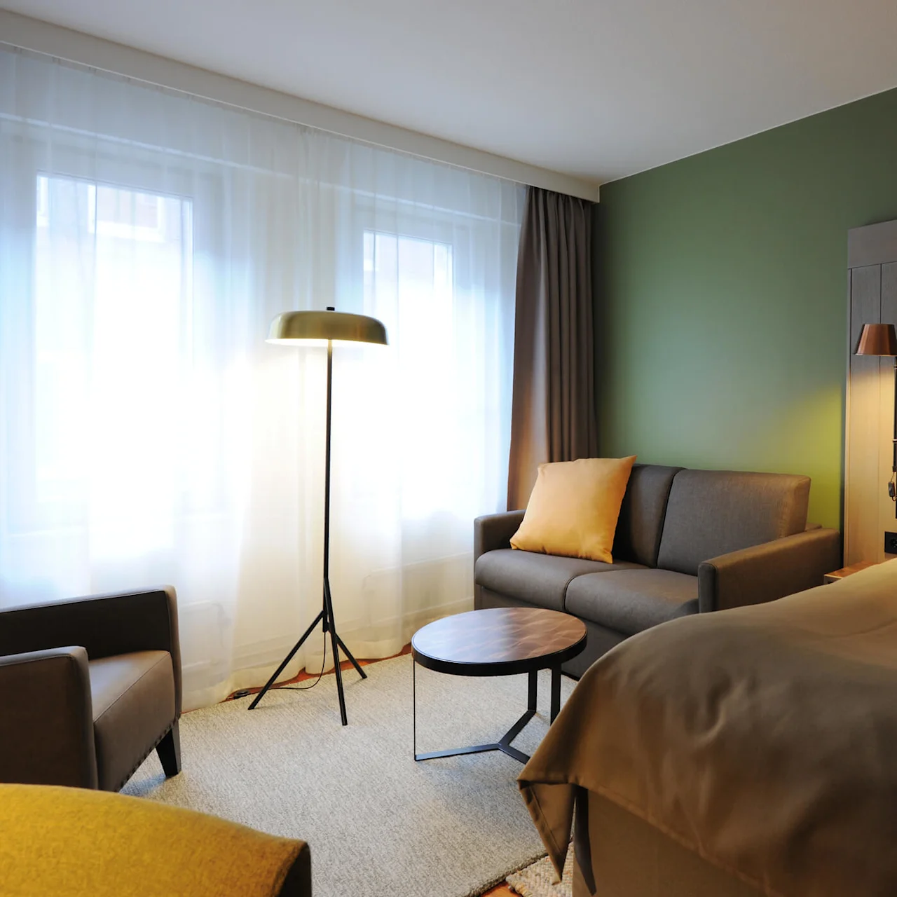 A cozy hotel room features a large bed with a wooden headboard, a sofa with cushions, and a floor lamp beside a window, creating a warm, inviting atmosphere.