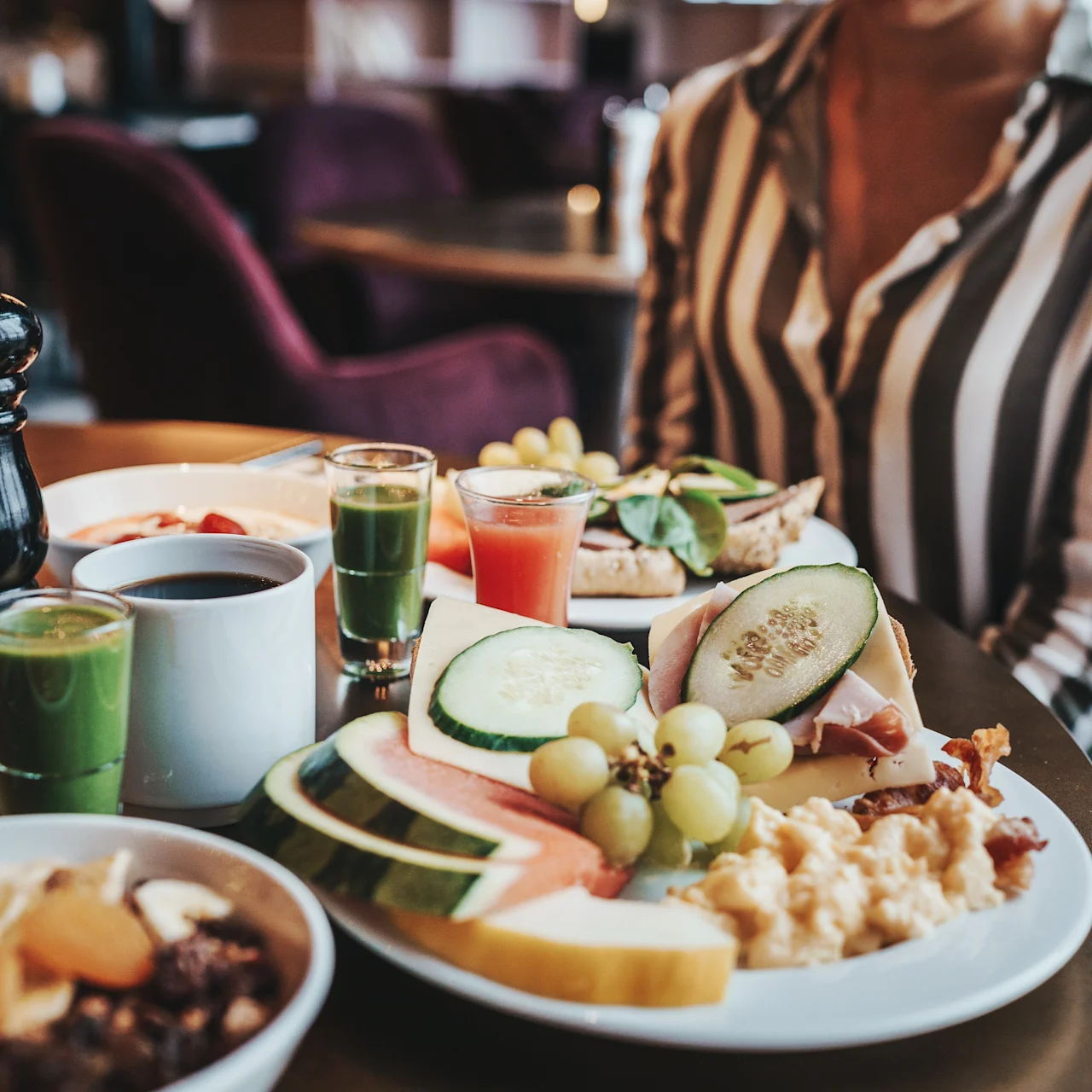 A breakfast table holds a plate with fruit and scrambled eggs, surrounded by juices and coffee. A person in a striped shirt sits nearby in a cozy dining setting.
