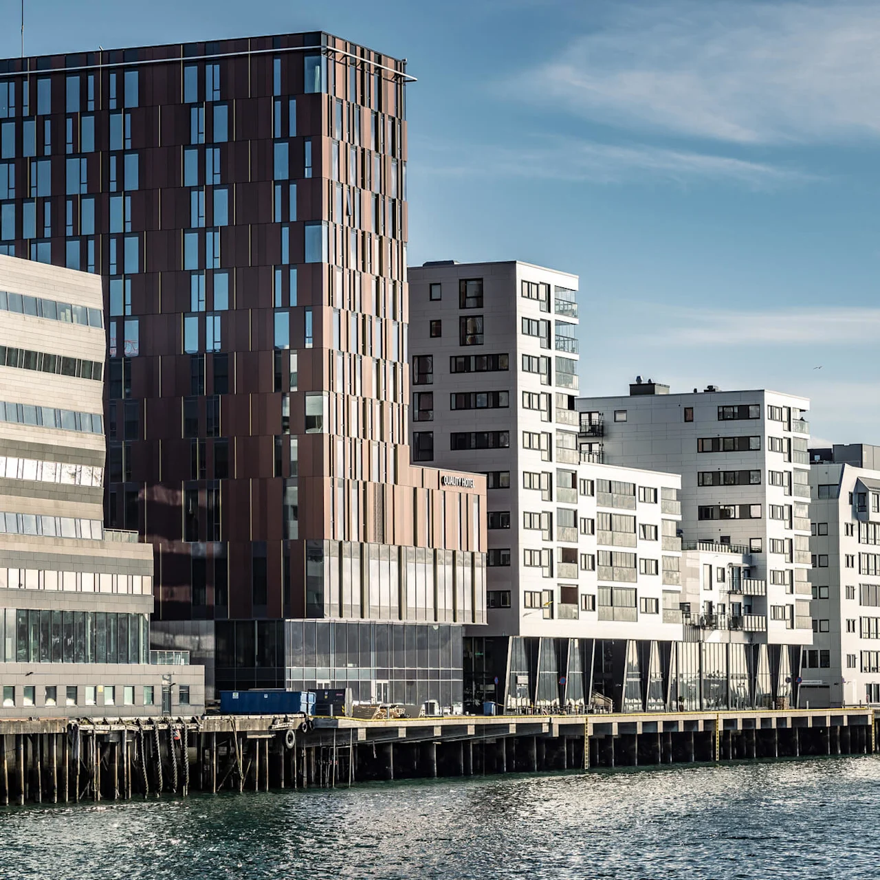 Modern buildings stand along a waterfront, reflecting sunlight. The largest structure is a brown, glass-panelled tower. The text "Radisson Blu" is visible on one building.