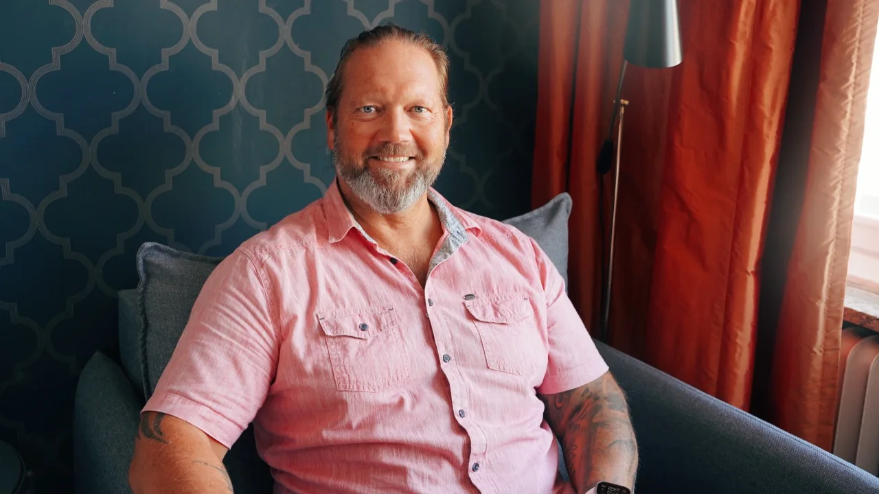 A smiling man sitting on a sofa, against a patterned wallpaper background; casual setting.