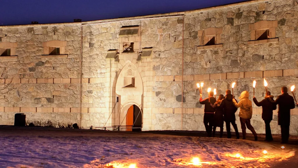 English: A group carrying torches walks on a snowy path before an illuminated stone fortress with arched entryway, under a deep blue evening sky. Trees stand bare nearby.