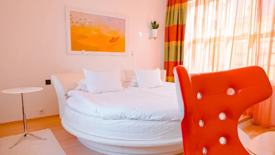 A round bed with white linens is in a brightly lit room, featuring colorful curtains and a modern orange chair. A painting and potted plant decorate the wall.