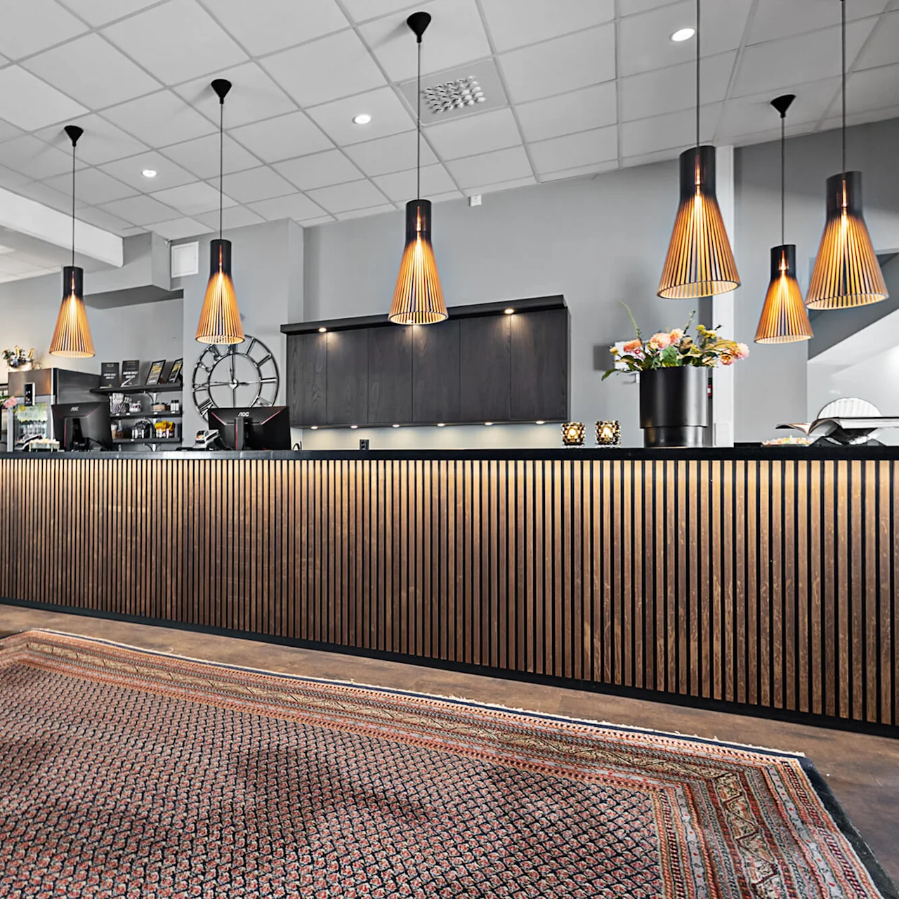 Ett modernt receptionsskrivbord med träpaneler och hänglampor i en stilfull lobby, med en mönstrad matta och dekorativa växter på disken.