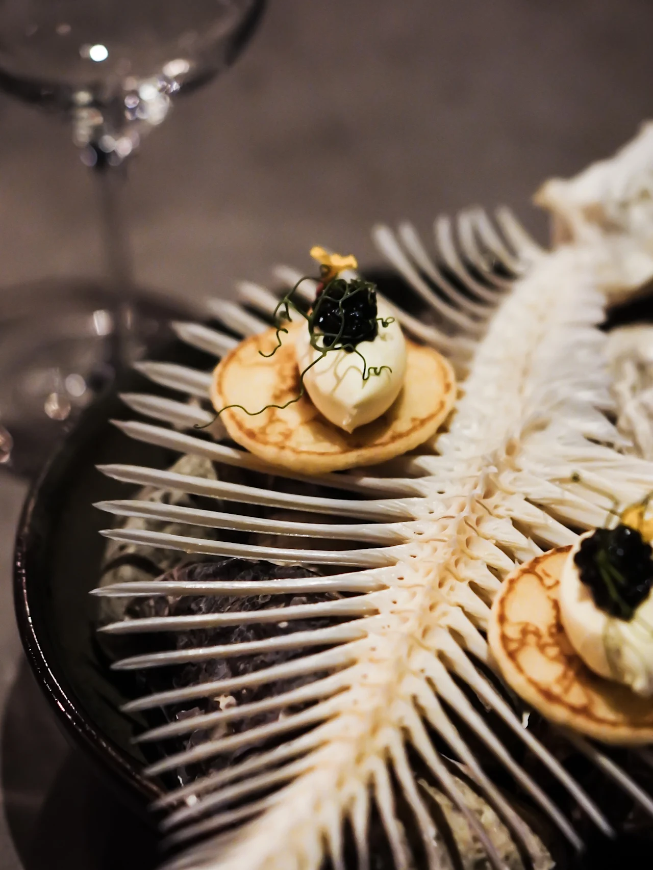 Elegant blinis med krem og kaviar, servert på en intrikat fiskebenstruktur, med et vinglass i nærheten.