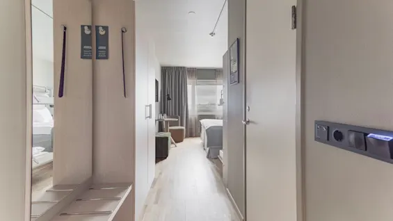A hotel room entrance with a mirror, coat rack, and "do not disturb" signs, leading to a bed and window.